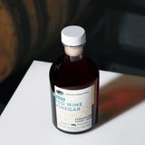 An angled, high-angle lifestyle shot of an 8.5 fluid ounce bottle of American Vinegar Works Ultimate Red Wine Vinegar sitting on a white surface, casting a deep red shadow with a wooden barrel visible in the dark background.