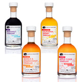 Four-bottle craft vinegar variety set on reflective surface — artisanal vinegar collection