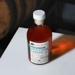 An angled lifestyle shot of an 8.5 fluid ounce bottle of American Vinegar Works Apple Cider Vinegar on a white surface, with a wooden barrel visible in the dark background.